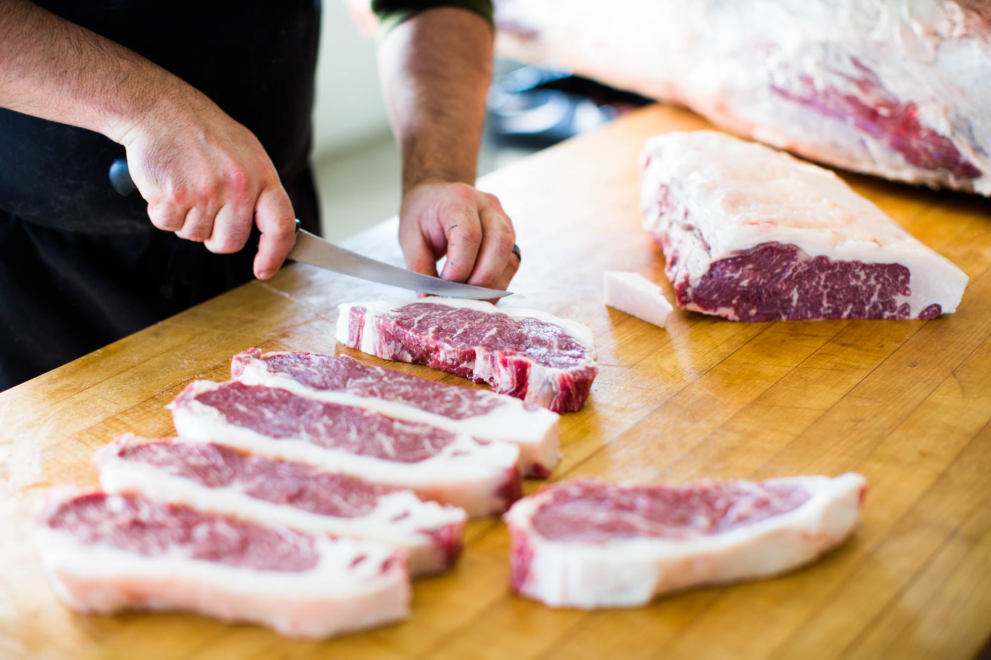 Butcher cutting a series of New York Strip Steaks on a butcher block.